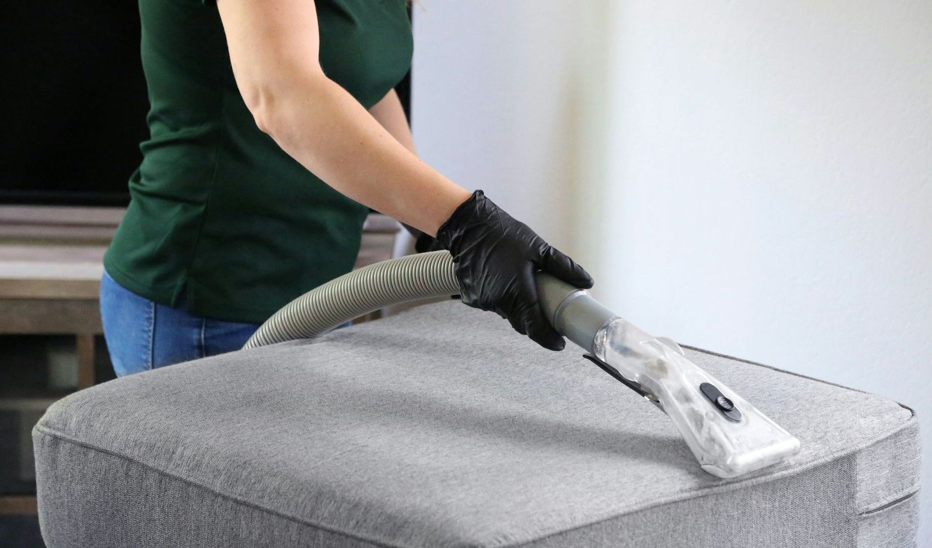 a woman in a green shirt and black gloves vacuuming a gray ottoman